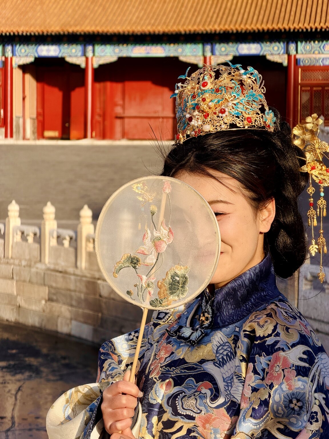 A woman in ornate traditional Chinese dress holding an embroidered round fan at the Forbidden City